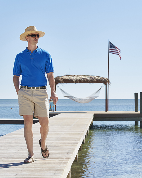 Man in a blue shirt and shorts walking on a dock with an American flag and hammock in the background.