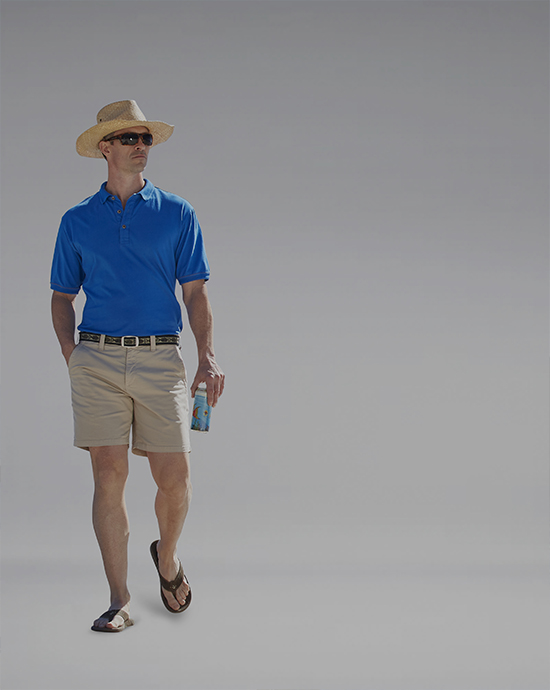Man in a straw hat, blue polo shirt, and khaki shorts, holding a drink, with a grey background.