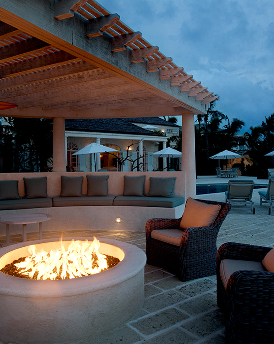 Evening view of an outdoor lounge with a fire pit, comfortable seating, and poolside background under a dim sky.