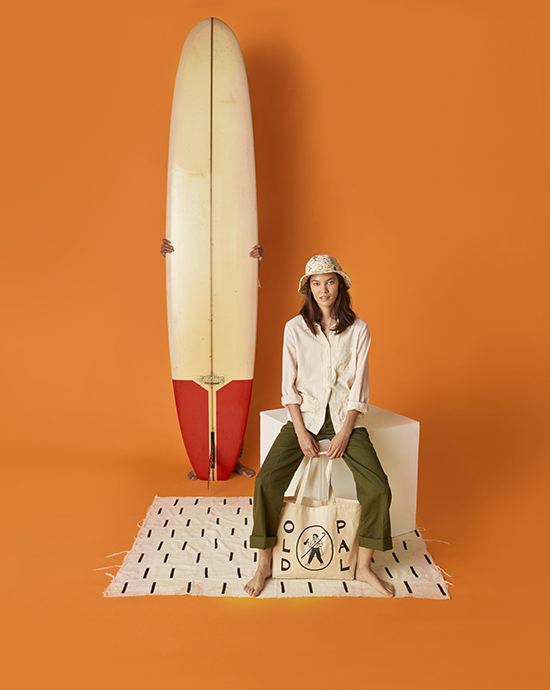 Background removal services for a woman sitting next to a surfboard against an orange background, dressed in casual summer attire.