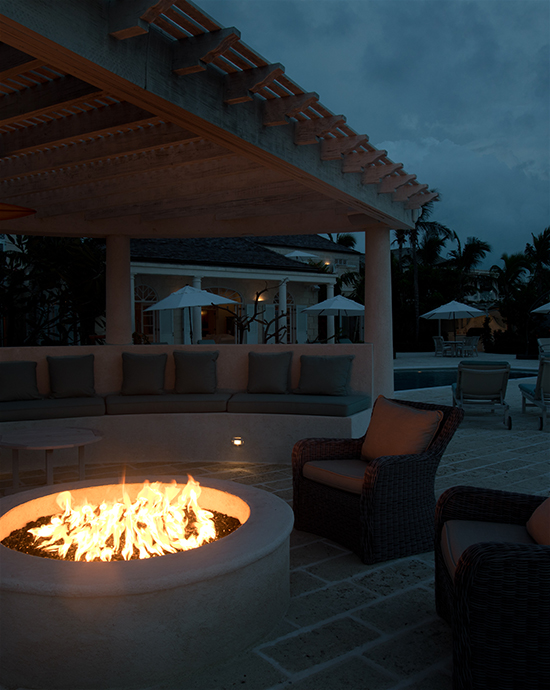 Outdoor patio with a lit fire pit at dusk, featuring cozy seating under a pergola and a dimly lit pool area in the background.