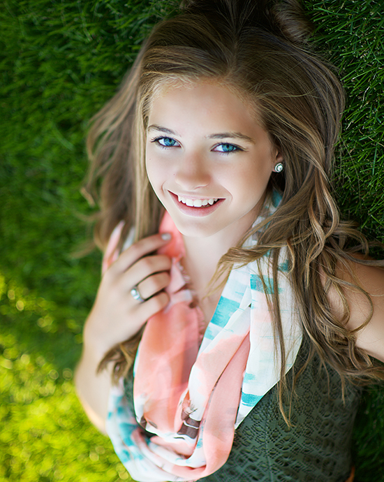 Young woman smiling while lying on grass, looking up at the camera with a bright and cheerful expression.