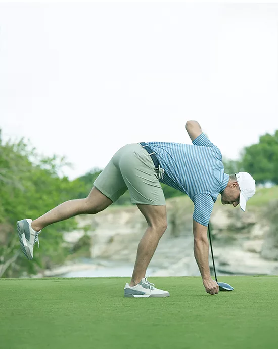 Man in a striped shirt and shorts bending to pick up a golf ball on a course.