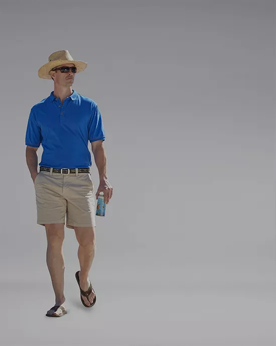 Man in a straw hat, blue polo shirt, and khaki shorts, holding a drink, with a grey background.