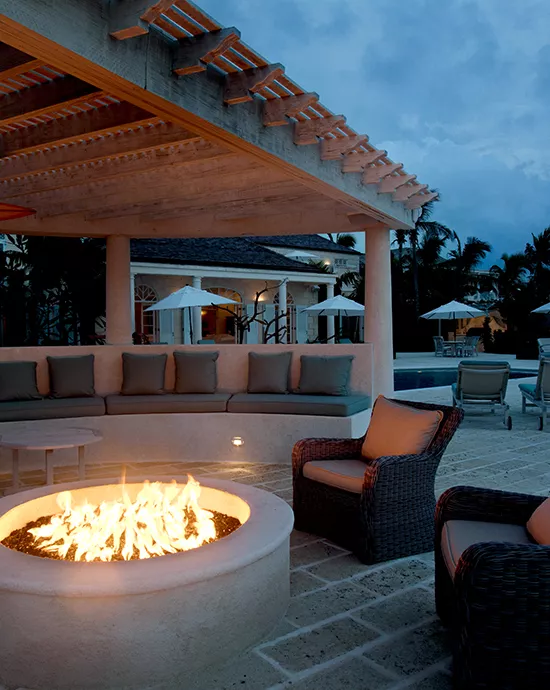 Evening view of an outdoor lounge with a fire pit, comfortable seating, and poolside background under a dim sky.