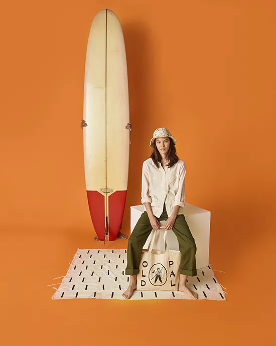 Background removal services for a woman sitting next to a surfboard against an orange background, dressed in casual summer attire.