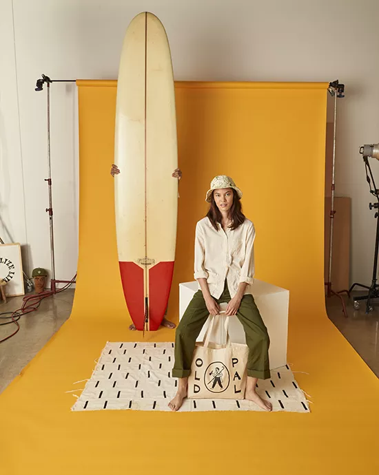 Background removal services for a woman sitting in front of a surfboard on a yellow backdrop, holding a tote bag.