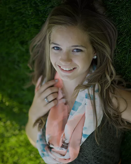 Young woman lying on grass, looking up and smiling, wearing a scarf and dark top.