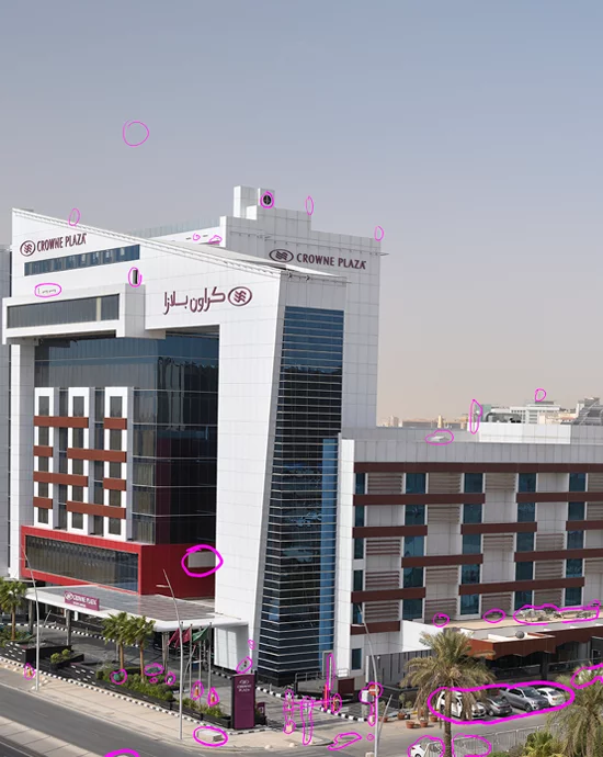 Modern hotel building with red and blue facade, "Crowne Plaza" sign, and Arabic script by busy street.