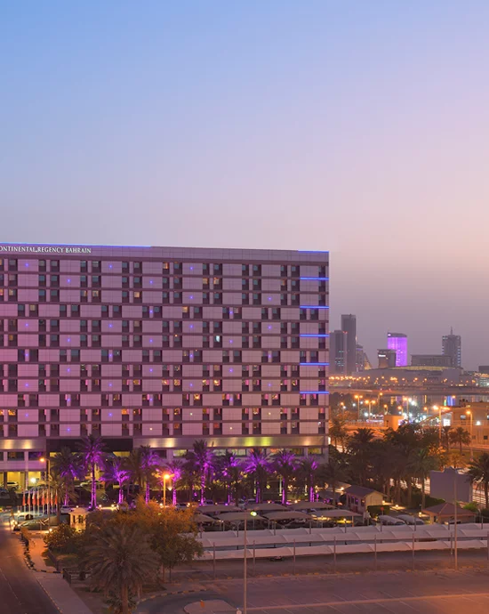 View of modern hotel facade at dusk with illuminated palm trees and city skyline.