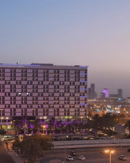 Dusk view of modern hotel facade with illuminated windows and city skyline background.
