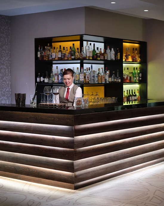Smiling bartender arranging glasses at modern bar with backlit liquor shelf.