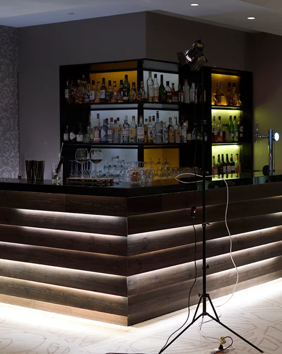 Dimly lit bar counter with bottles, glasses, and spotlight in front.