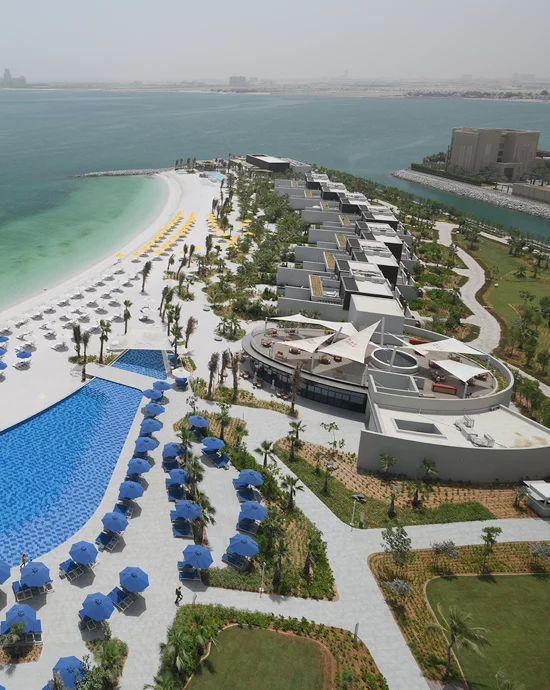 Aerial view of luxurious beachside resort with pools, umbrellas, and modern architecture along vibrant coastline.