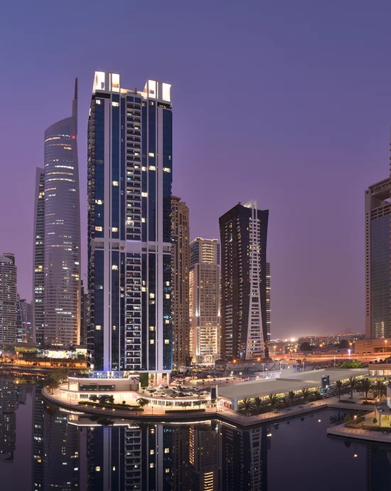 Twilight view of modern city waterfront with illuminated skyscrapers reflecting in river.