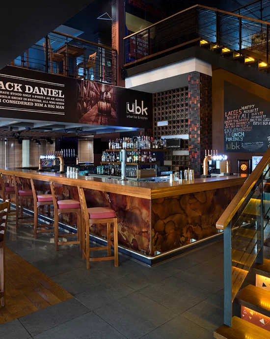 Modern bar interior with wooden stools, large liquor selection, and urban-themed decor. Promotional banners above.