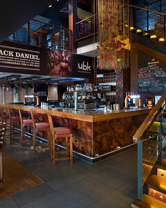 Modern bar interior with wooden accents, well-stocked bar, and vibrant lighting. Promotional banners above.