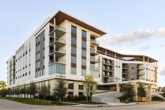 Modern multi-story residential building with balconies, landscaped greenery, and a curved driveway.