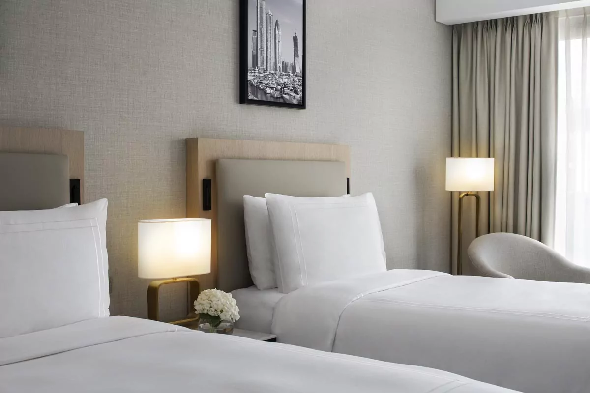 Bright hotel room with two white beds, modern lamp, armchair, and cityscape photo for Real Estate Photo Editing Services.