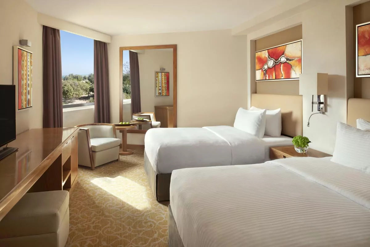 Bright hotel room with two beds, modern decor, flat-screen TV, and large windows for Real Estate Photo Editing Services.